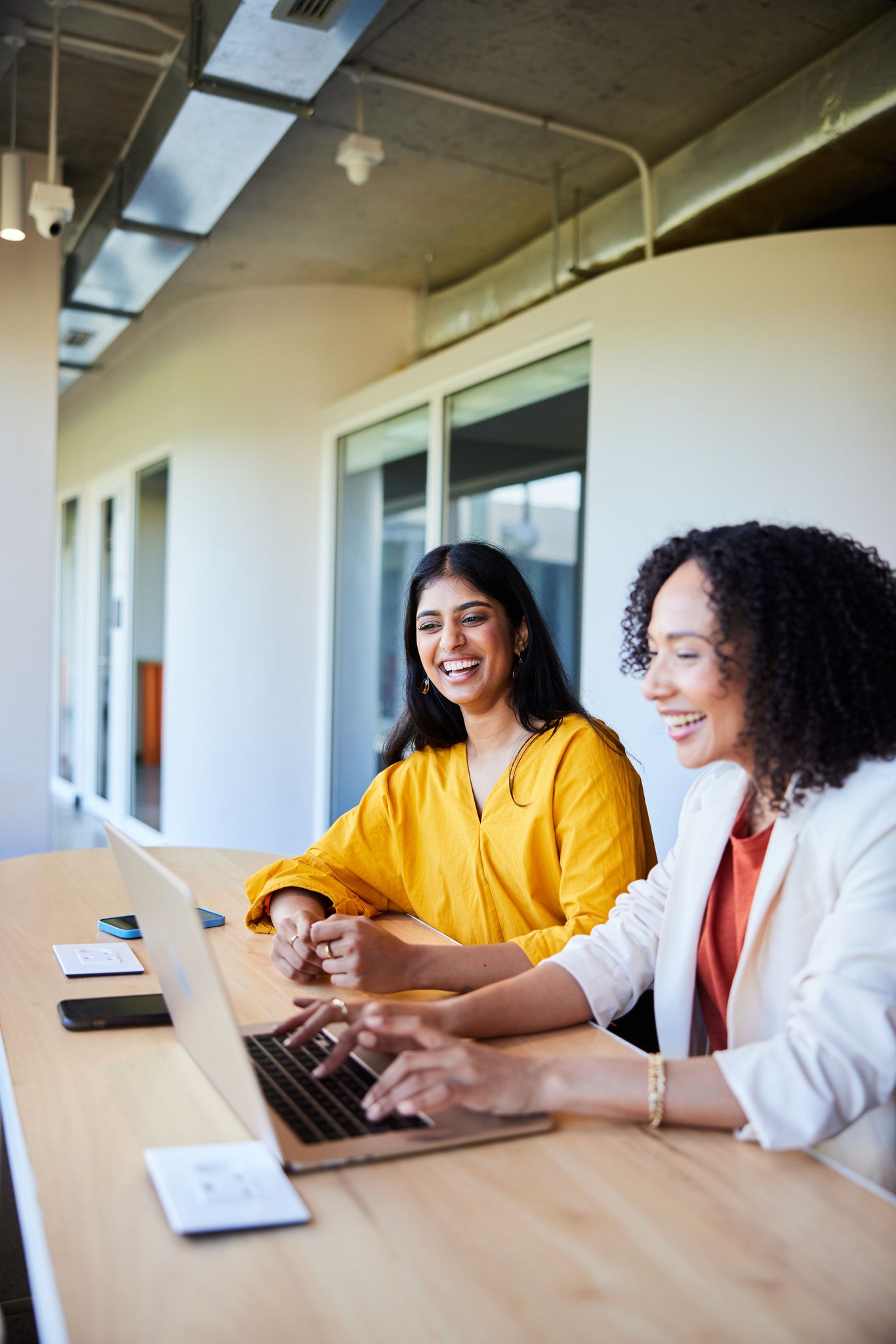 Two young businesswoman smiling and talking together while working on a laptop in an office lounge area