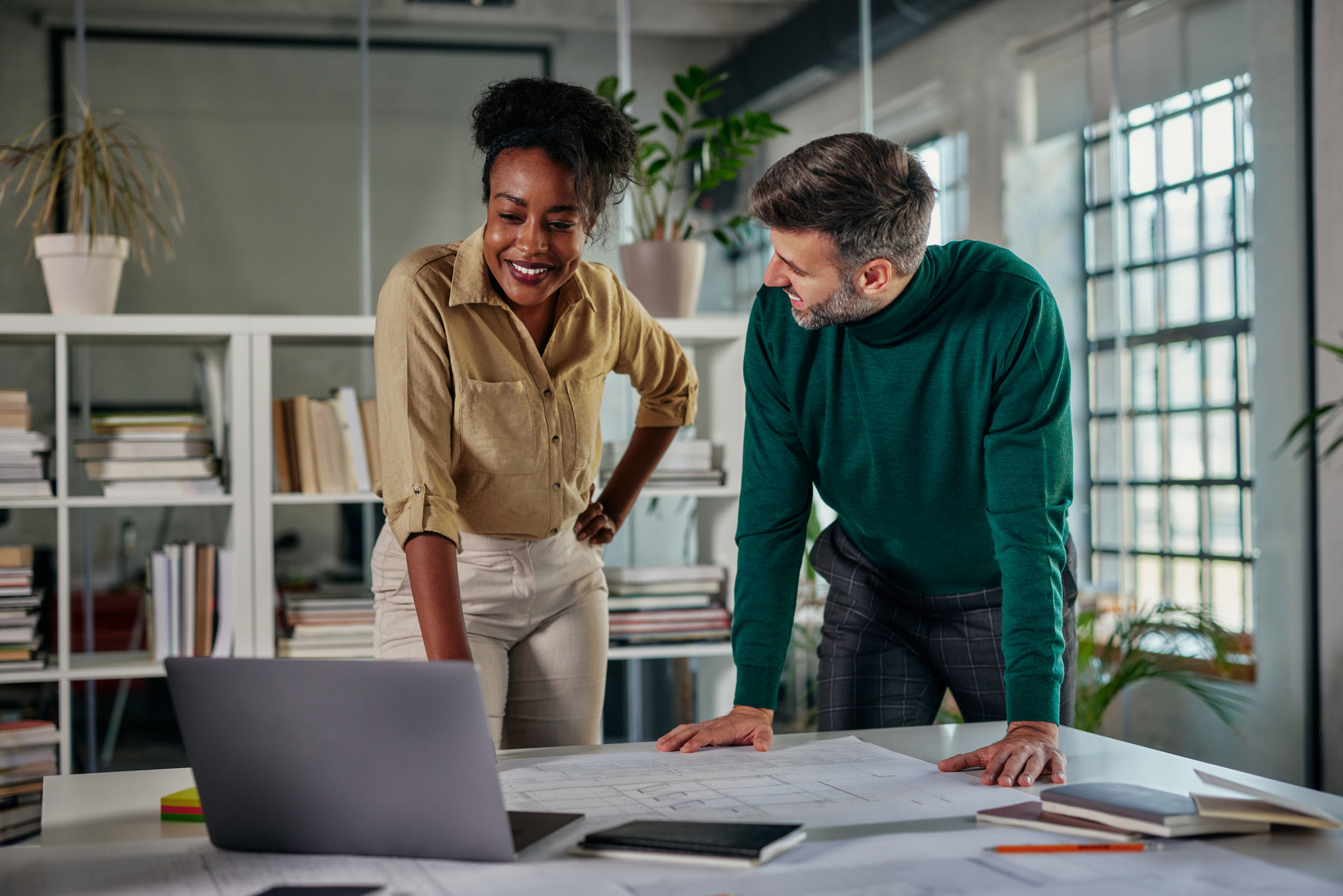 African businesswoman with laptop explaining project details to male caucasian colleague at office desk. Business partners having meeting in coworking office space.