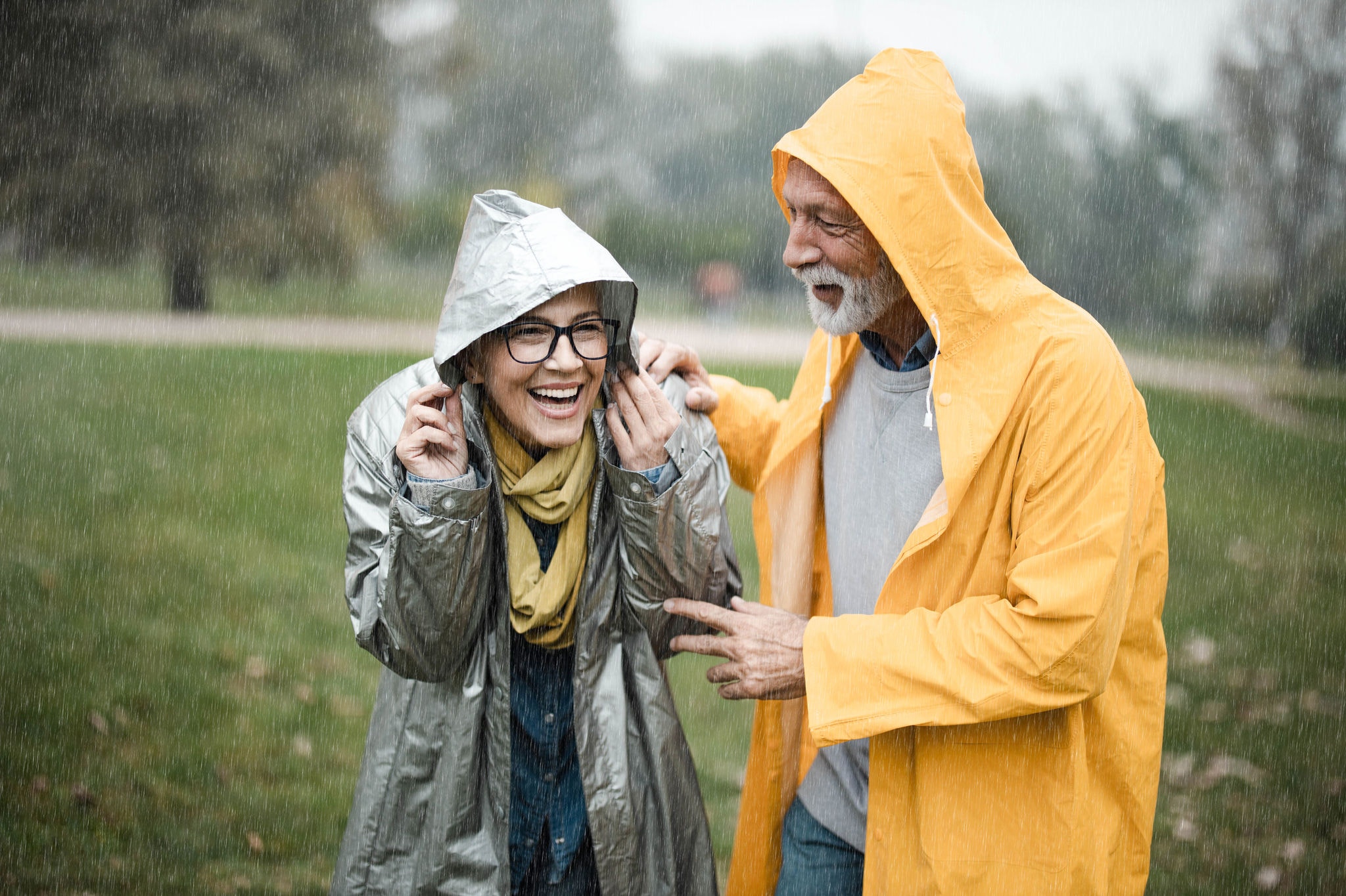 Happy couple in raincoats covering their heads during rainy day in nature.