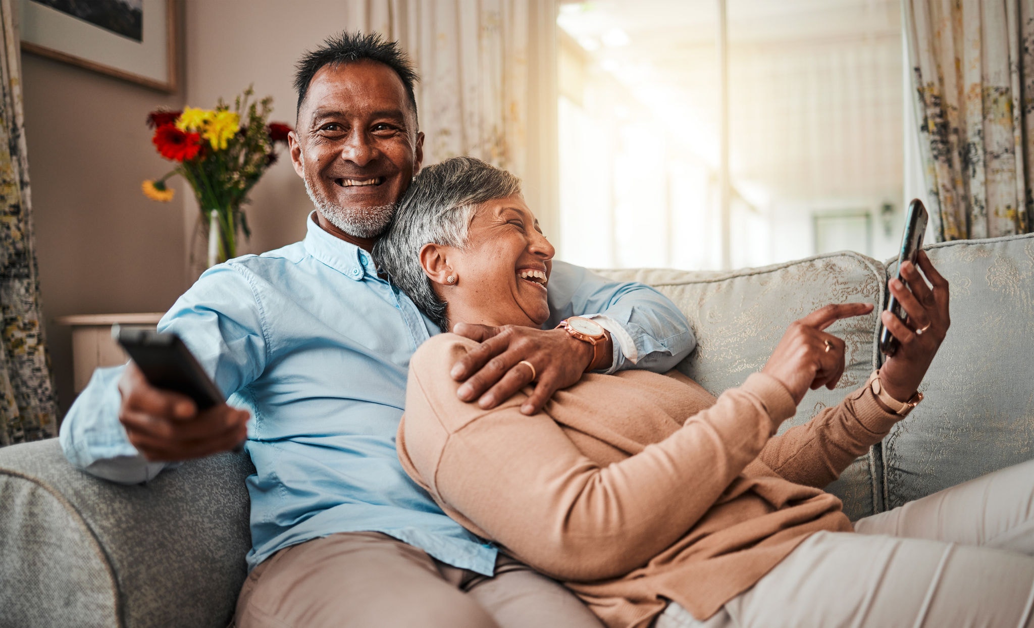 Senior couple relaxing on the couch, watching TV while also using their phones