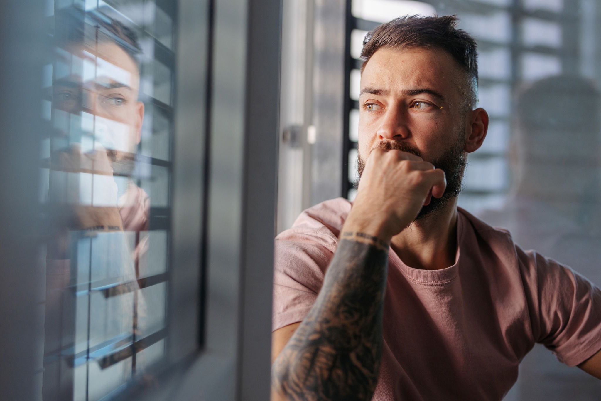 Calm man in pink t-shirt with tattoo on arms looking through window