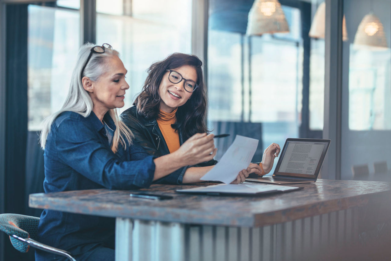 Two women analyzing documents while sitting on a table in office. Woman executives at work in office discussing some paperwork.